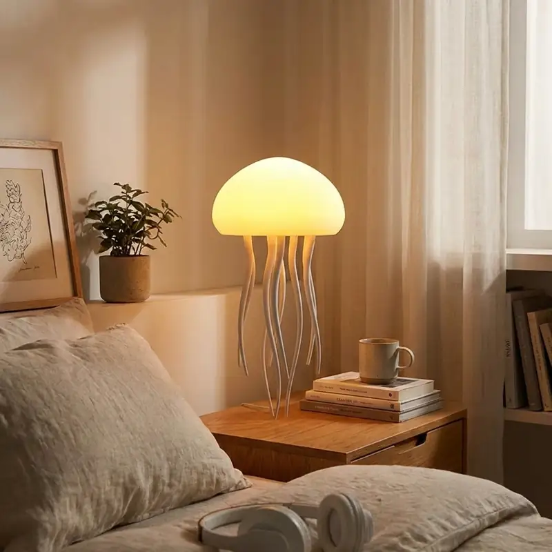 A jellyfish-shaped table lamp with a glowing warm yellow dome and slender white tentacles sits on a wooden nightstand. The scene is a cozy, modern bedroom with soft linen bedding, a stack of books, a ceramic mug, and a small potted plant against a beige wall, creating a peaceful and healing atmosphere.