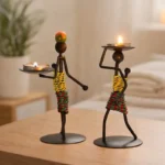 Two human-shaped metal candle holder figurines holding lit tea lights, minimalist iron design with colorful wrapped bodies, placed on a wooden table in a calm modern home setting.