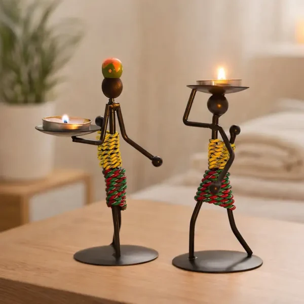 Two human-shaped metal candle holder figurines holding lit tea lights, minimalist iron design with colorful wrapped bodies, placed on a wooden table in a calm modern home setting.