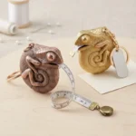 Chameleon-shaped metal keychain measuring tapes in gold and rose gold finishes with retractable metric rulers extended, placed on a clean tabletop with everyday items.