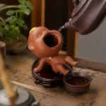 Funny purple clay tea pet in baby boy shape peeing water into a tea cup.