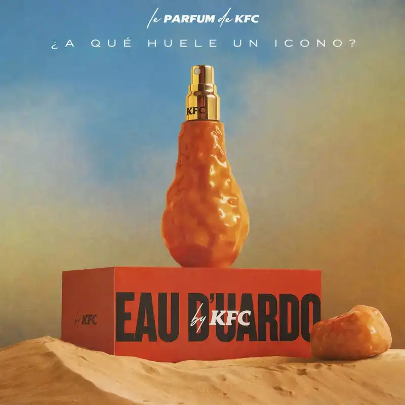 A close-up of the KFC Eau Duardo fragrance, a novelty perfume shaped like a fried chicken drumstick with a golden spray top and the KFC logo, placed on a red box labeled ‘EAU D’UARDO by KFC’ against a warm gradient background.
