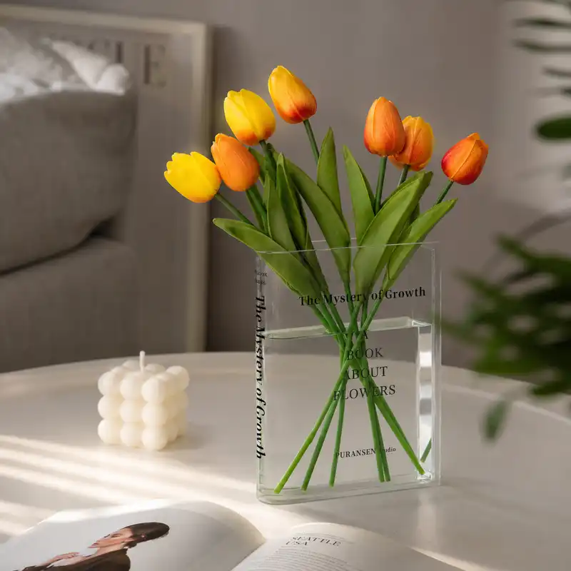 Clear acrylic book vase with yellow and orange tulips displayed on a white bookshelf beside design books in a modern home interior.