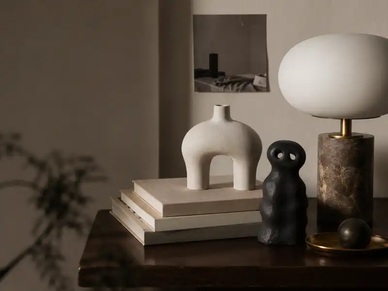 A warm, minimalist editorial still life featuring sculptural decorative objects on a wooden desk, softly lit in neutral tones, evoking a calm and slightly surreal atmosphere about attraction to unusual objects in modern home design.