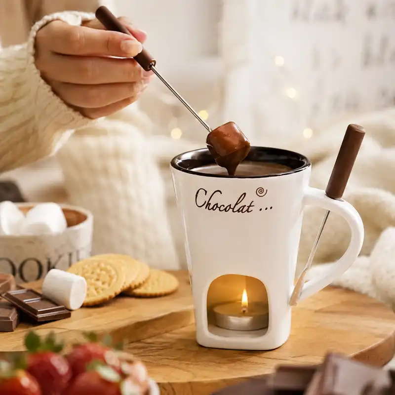 A white chocolate fondue mug with a tealight candle underneath, melted chocolate inside, and a dipping fork holding a marshmallow.